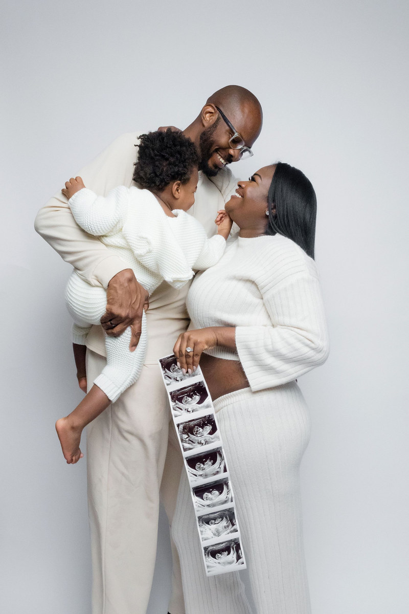 Couple in white lifting a toddler while holding a strip of ultrasound photos