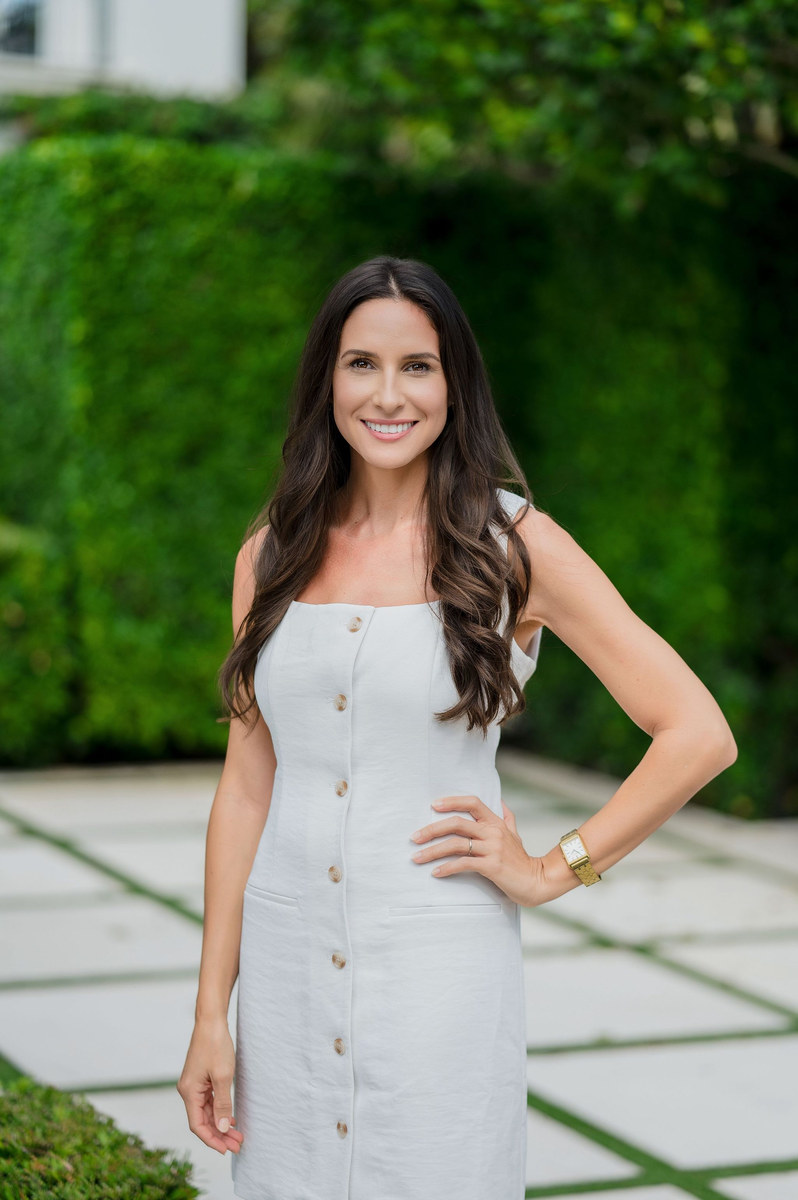 Woman in a white sleeveless dress standing on a garden walkway, hand on her hip