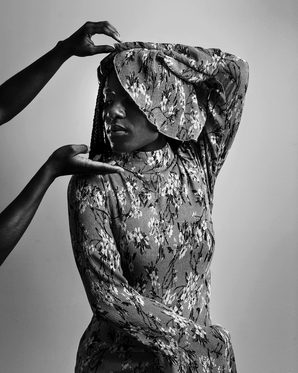 Black and white portrait of a woman in a floral dress and hat, with hands reaching in from outside the frame
