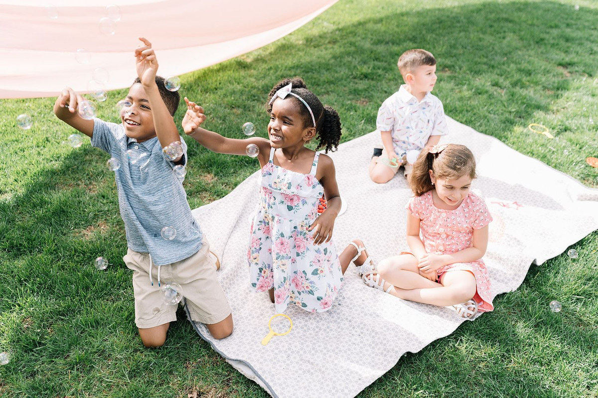 Three children playing on a picnic blanket in a grassy yard