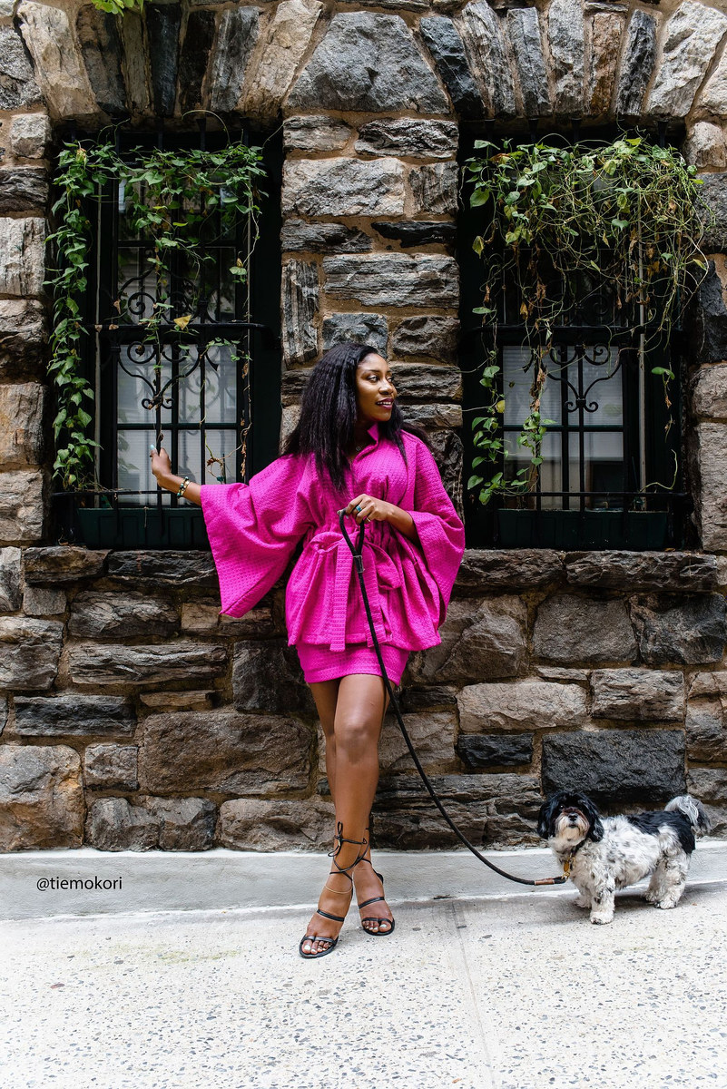 Woman in a pink dress walking a small dog past a stone building