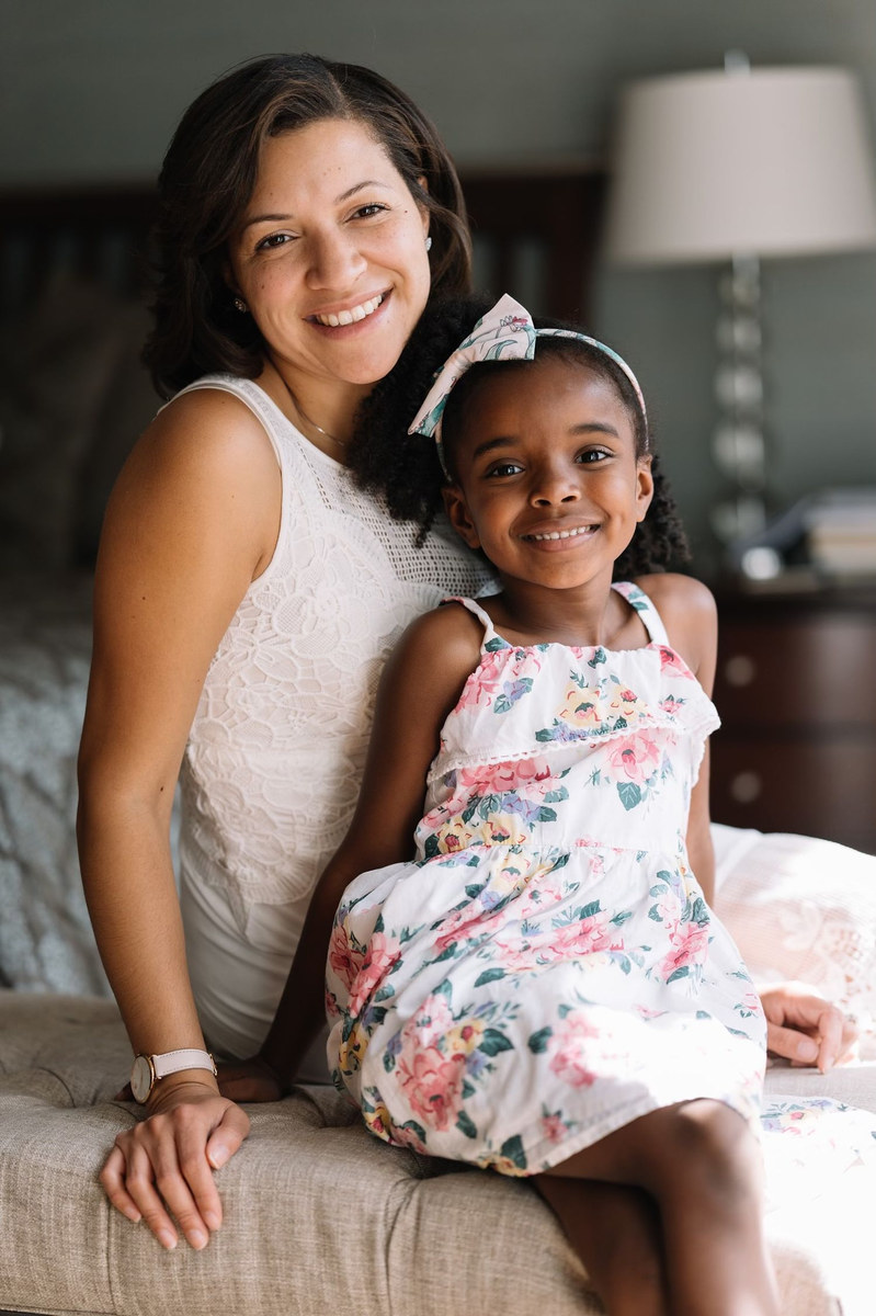 Woman and young girl in a floral dress smiling together, seated on a bed
