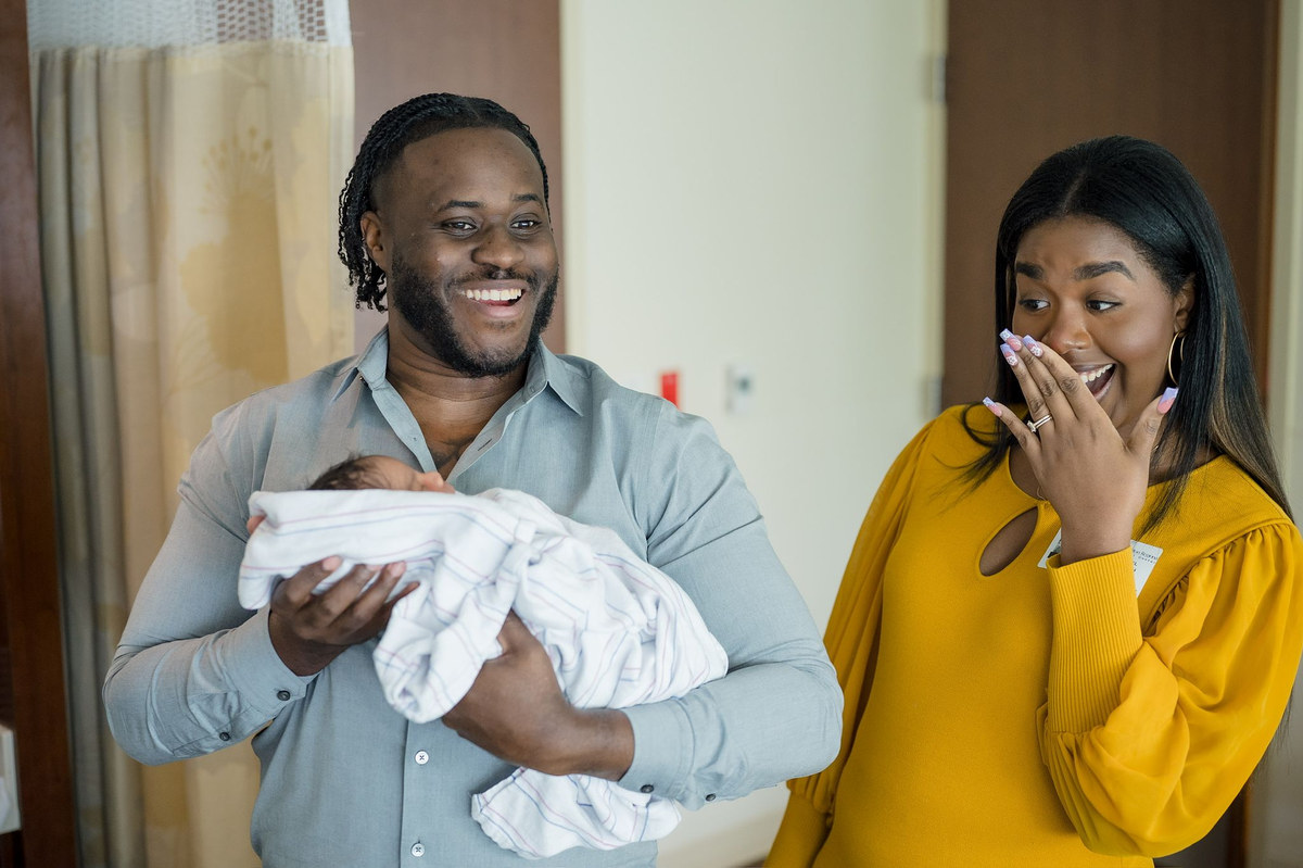 Man holding a newborn wrapped in a blanket as a woman beside him laughs