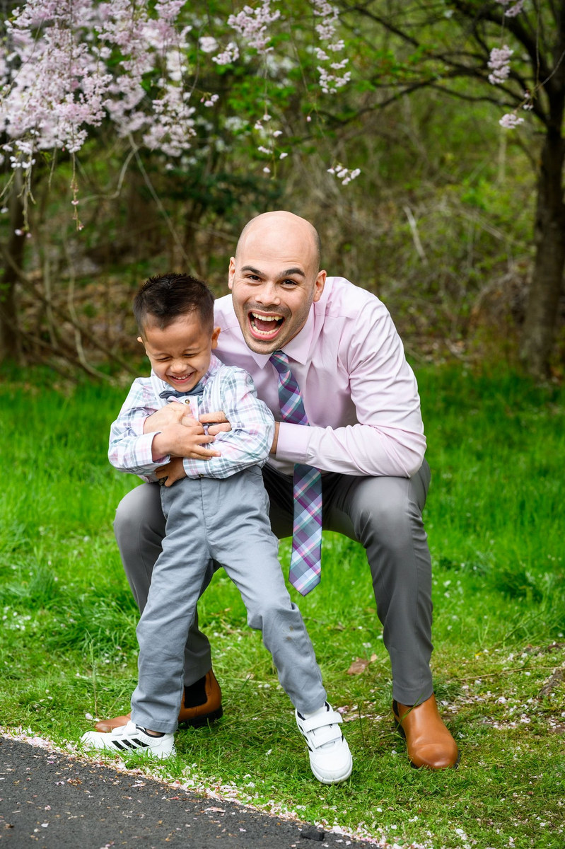 Man crouching beside a laughing toddler on the grass under a blossoming tree